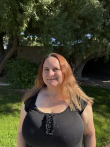 Kaila Allen, A woman with long, reddish-blonde hair is standing outdoors in natural light, smiling at the camera. She is wearing a black top with a scoop neckline and a light-colored cardigan. The background is green, likely trees or bushes, indicating she is in a park or garden setting.